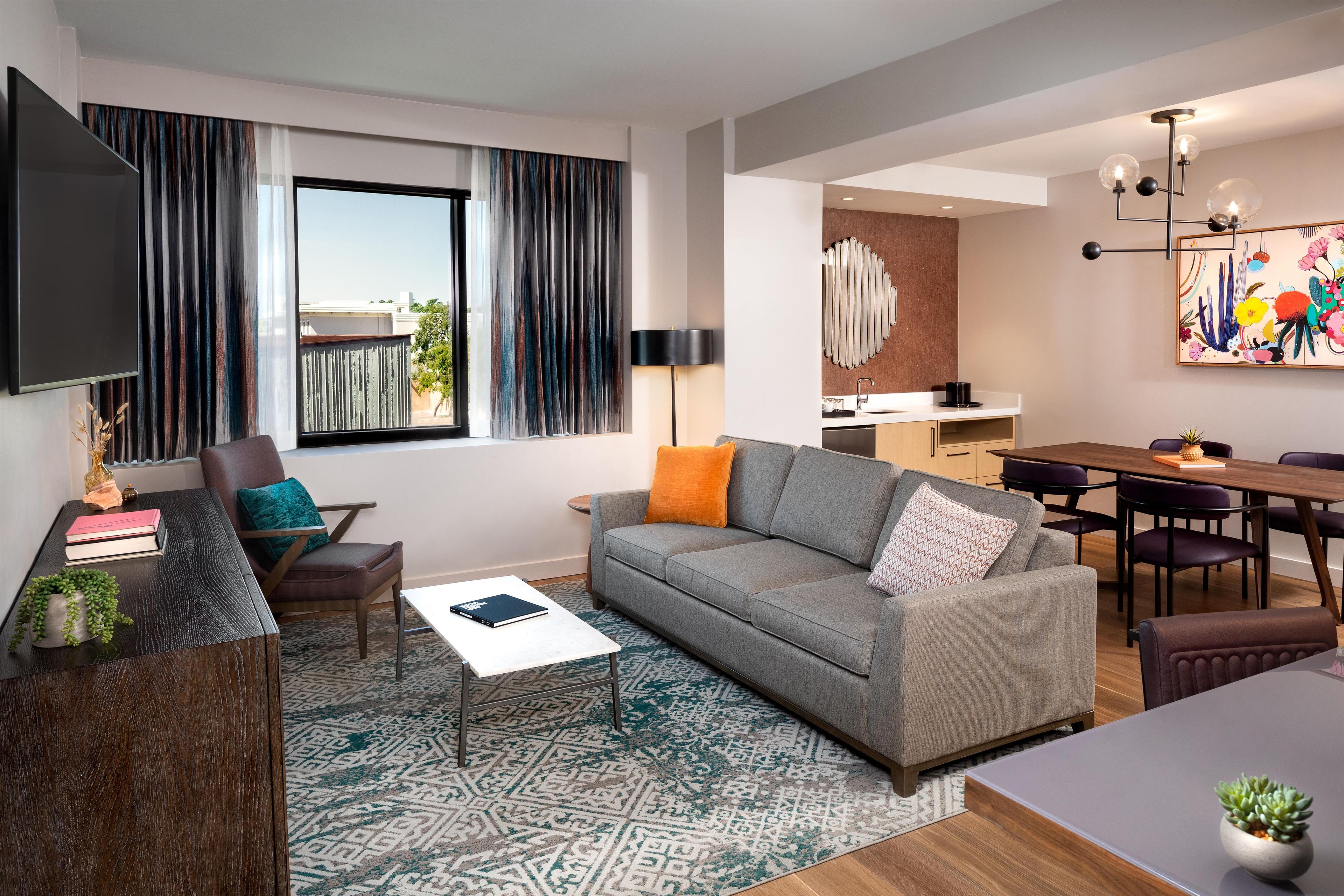 Una sala moderna con un sofá gris, una mesa de centro y un televisor. Hay una zona de comedor con una mesa y sillas, y una cocina al fondo.