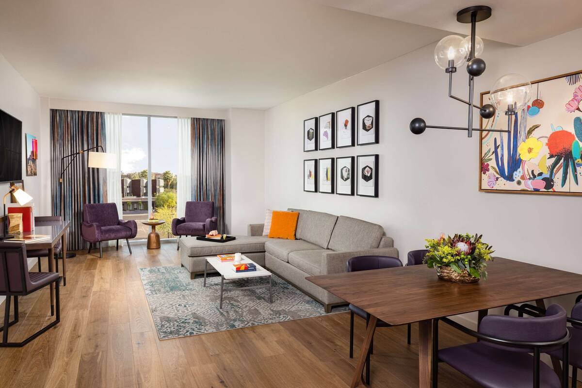 Una sala de estar moderna con un sofá, sillas, una mesa de centro y una mesa de comedor. Hay cuadros enmarcados en la pared y un cuadro de colores.