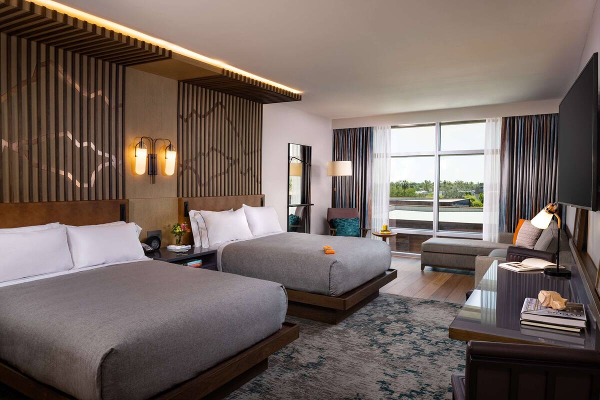 Habitación de hotel con dos camas, un escritorio, una silla, un ventanal y vistas al exterior.