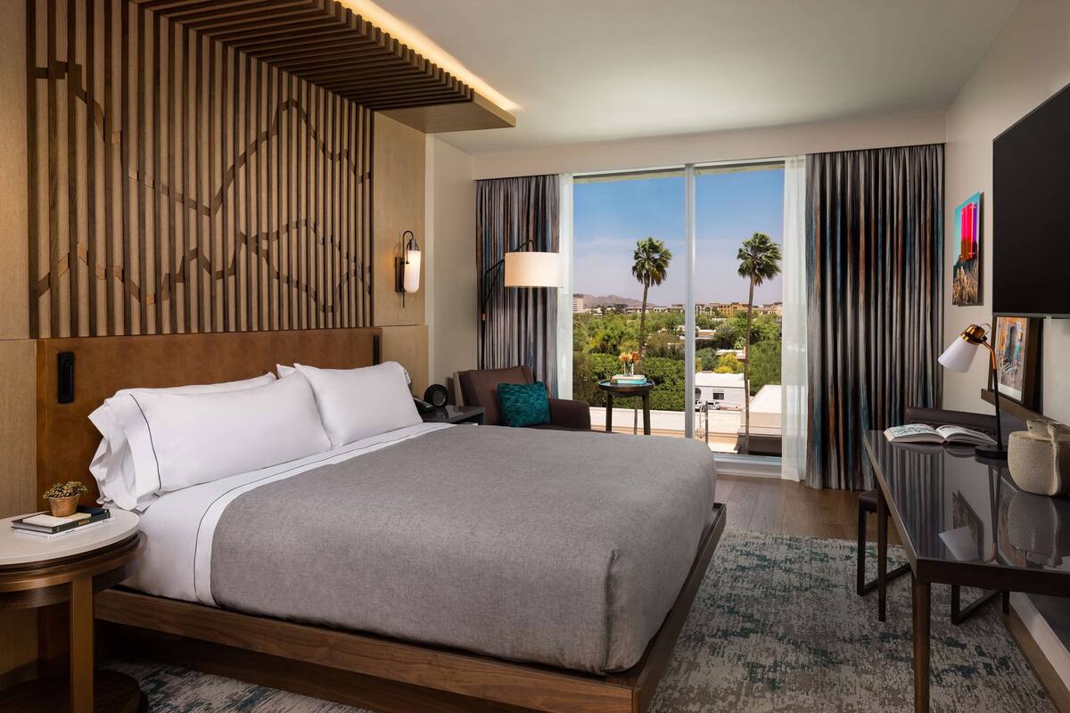 Habitación de hotel moderna con una cama grande, un escritorio y vistas a palmeras y edificios.