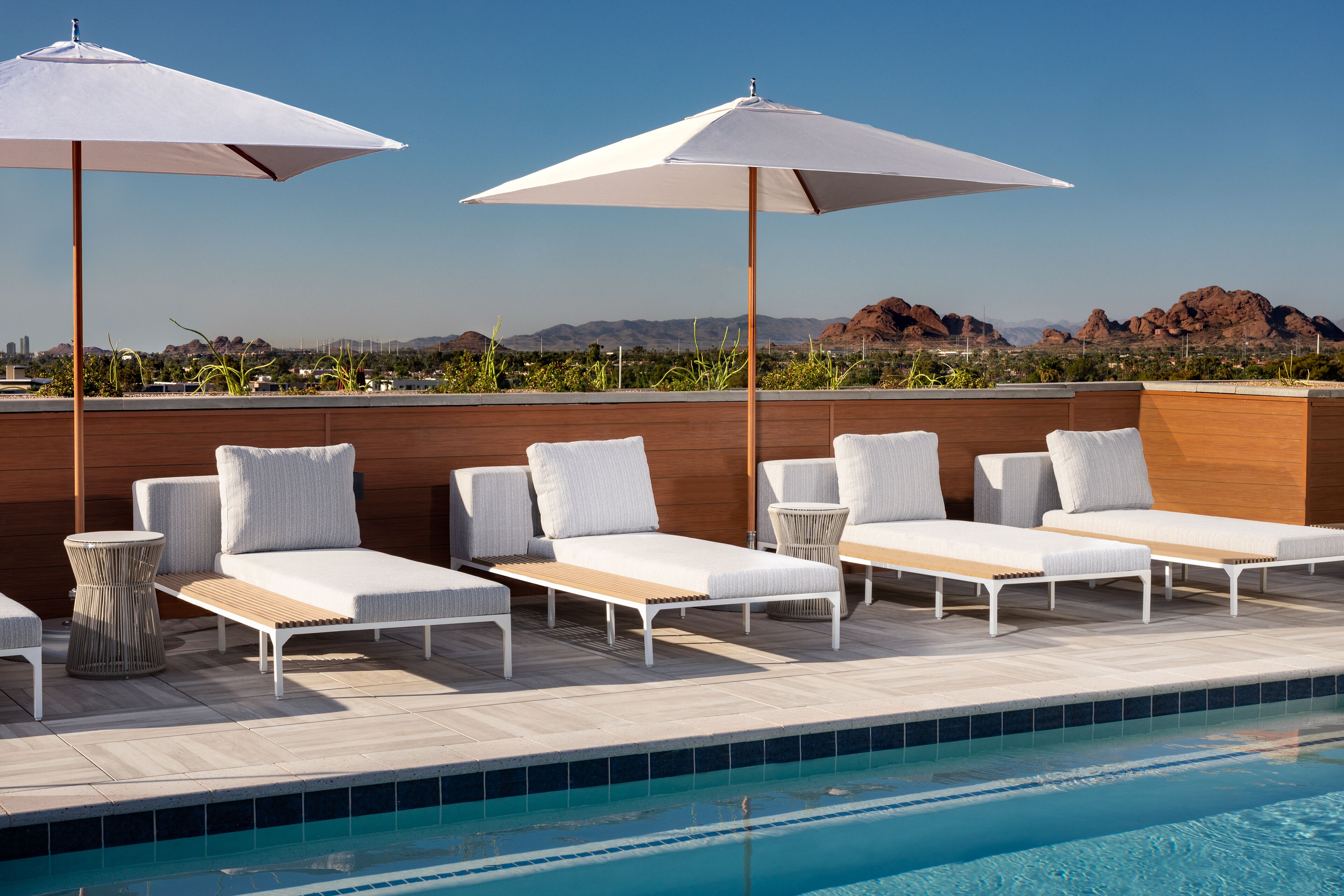 A rooftop pool area with lounge chairs and umbrellas, a wooden deck, and a view of distant mountains.