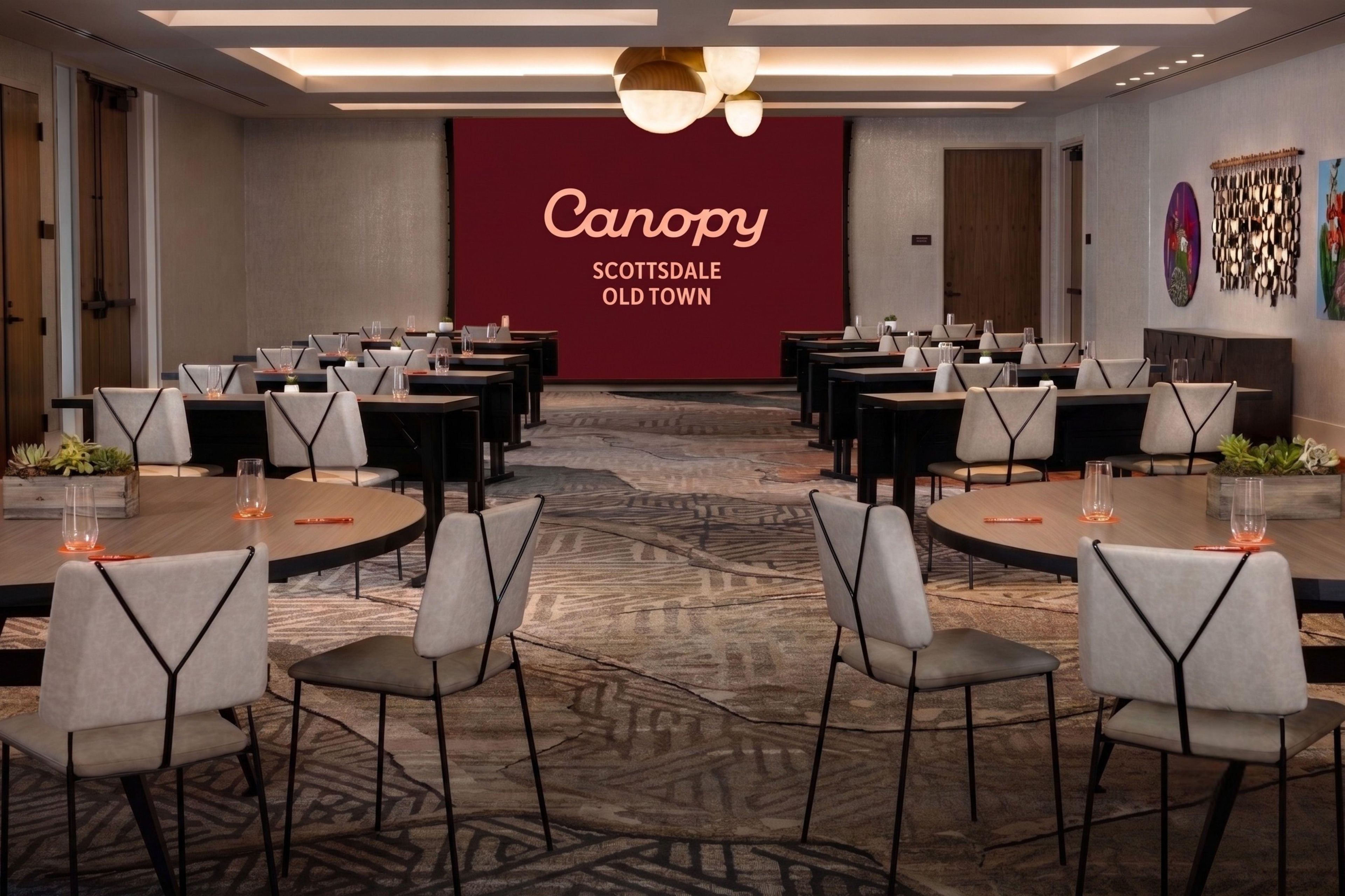 A conference room with tables and chairs arranged for a meeting, a large screen displaying 'Canopy Scottsdale Old Town', and a modern ceiling design.