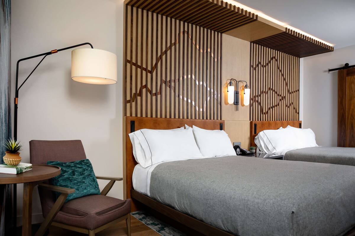 Habitación de hotel con dos camas, un escritorio con una silla, una lámpara y un panel decorativo en la pared.