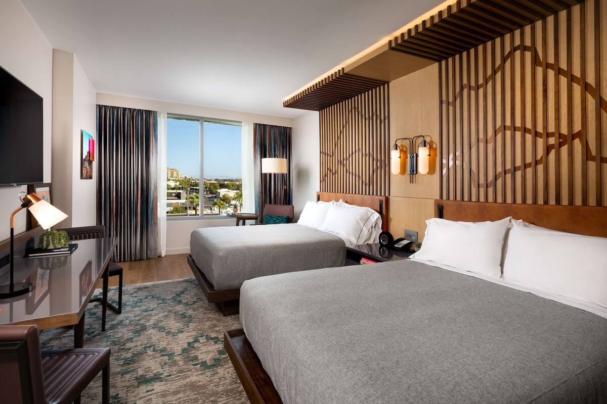 Habitación de hotel con dos camas, un escritorio con lámpara, televisión y vista a la ciudad.