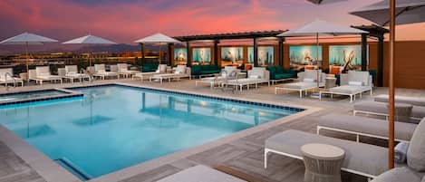 A rooftop pool area with lounge chairs and umbrellas, a pool, and a cityscape view at dusk.