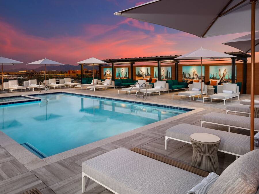 A rooftop pool area with lounge chairs and umbrellas, a pool, and a cityscape view at dusk.