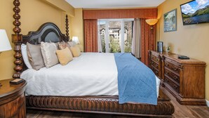 A bedroom with a large bed, wooden headboard, a dresser, a television, and a view of a building through the window. - Platinum Luxury Condo in Heart of Avon A606 (Avon)