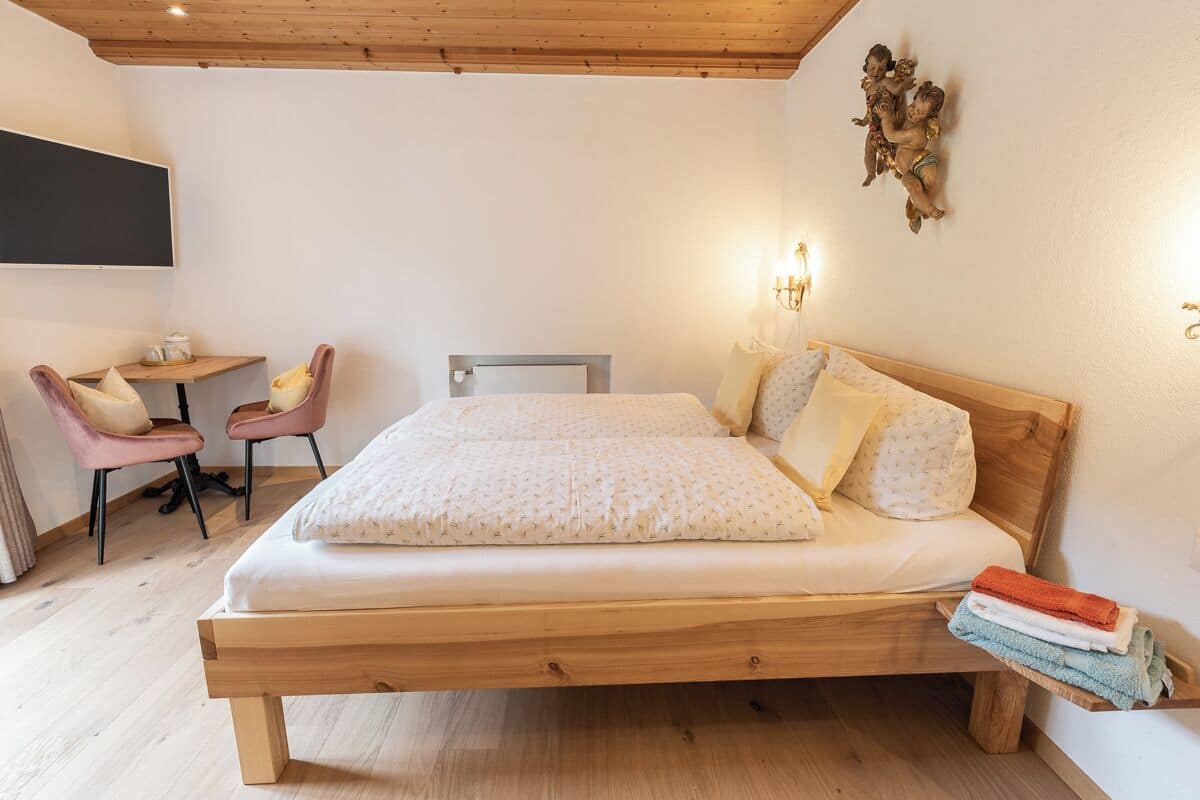 A bedroom with a wooden bed, two chairs, a small table, and a wall-mounted TV.