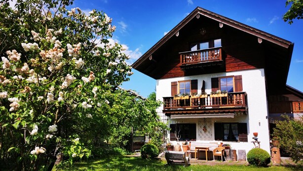 Un chalet de dos pisos con balcón, rodeado de exuberante vegetación y árboles en flor.