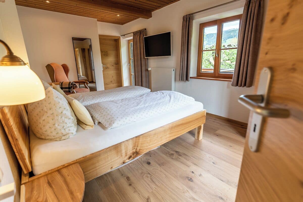 A bedroom with a wooden bed, a TV, a chair, and a window with curtains.
