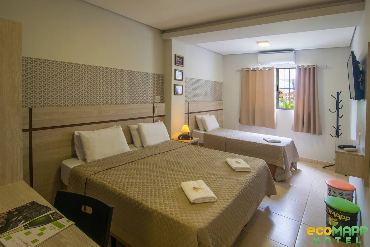 Ein Hotelzimmer mit einem großen Bett, einem kleineren Bett, einem Schreibtisch mit Stuhl, einer Lampe und einem Fenster mit Vorhängen.