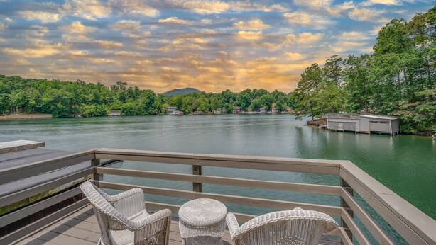 A wooden deck with wicker chairs overlooking a calm river, surrounded by lush greenery and a clear sky.
