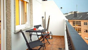 Un balcón con mesa y sillas, una ventana con cortinas, y vistas a edificios al fondo.