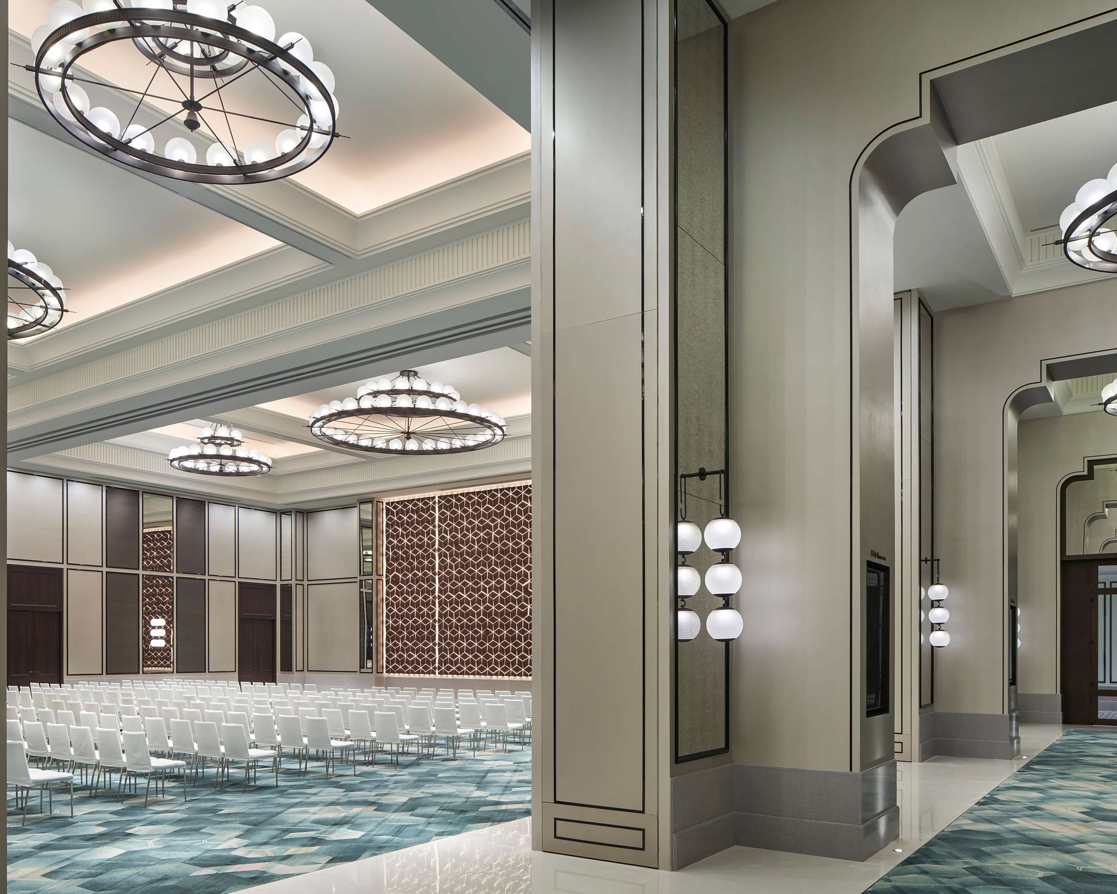 A spacious conference room with rows of white chairs, large chandeliers, and a patterned wall.