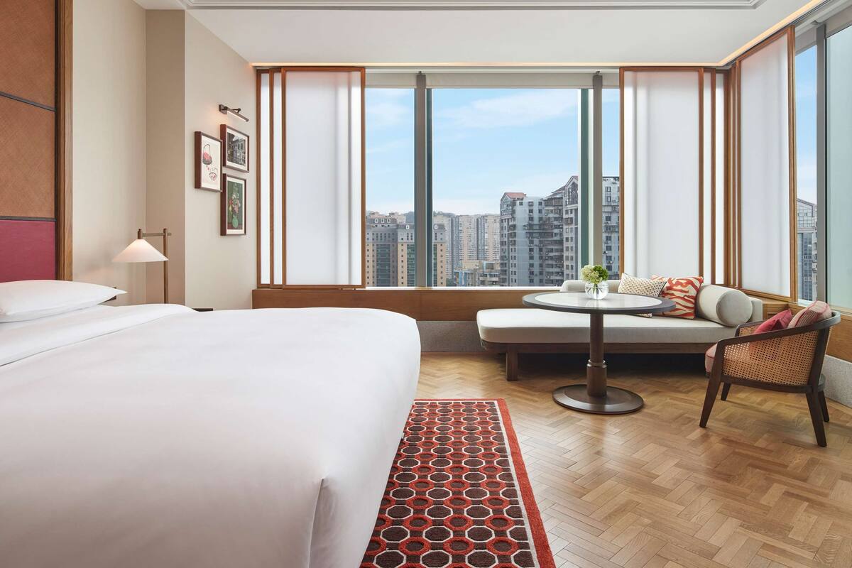 A hotel room with a large bed, a sofa, a small table, and a view of city buildings.