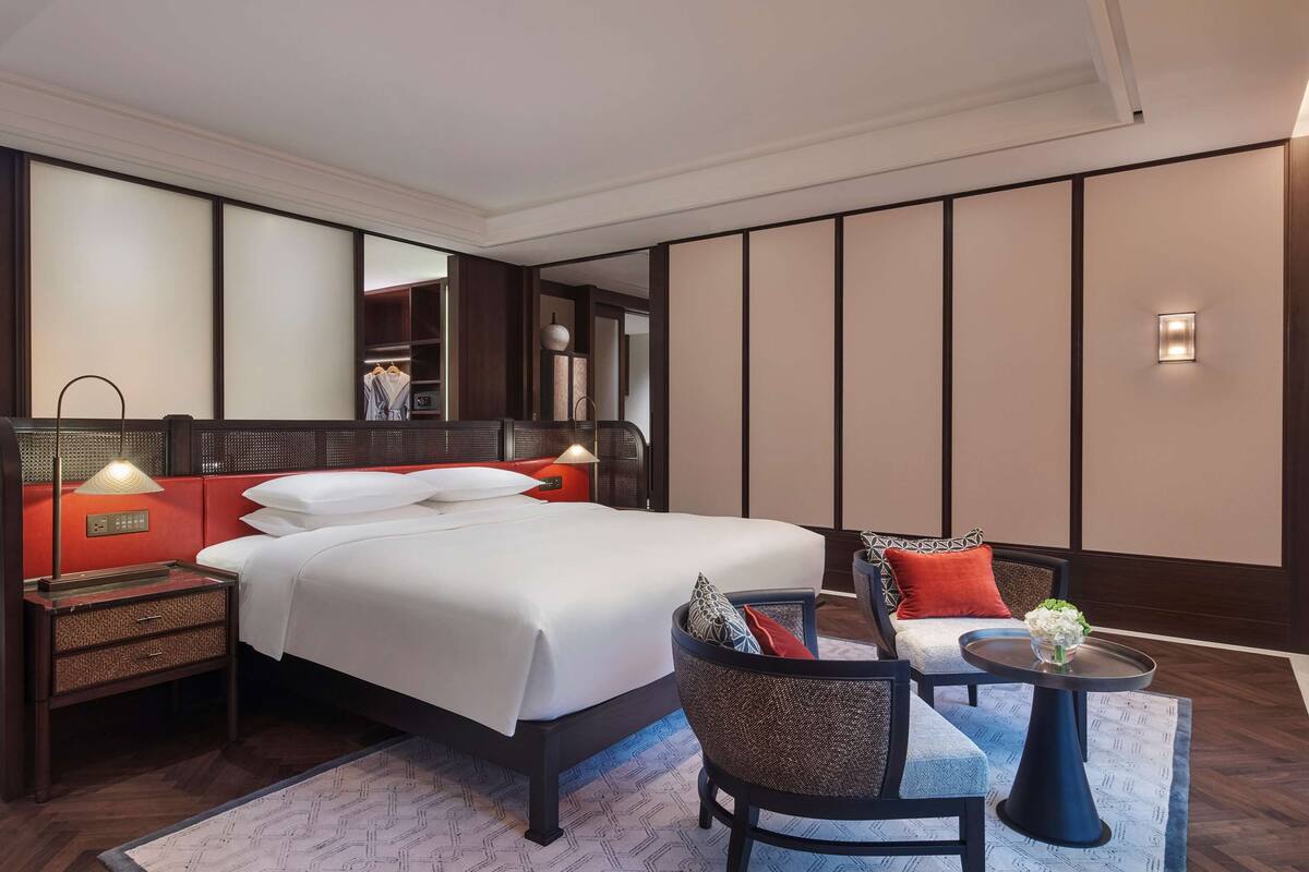 A hotel room with a large bed, a bedside table with a lamp, a chair, a small table with a vase, and a mirror on the wall.