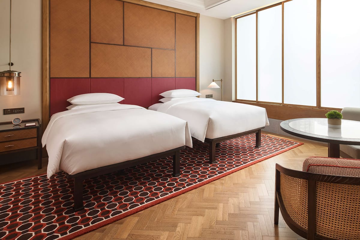 A hotel room with two beds, a round table, a chair, a nightstand, and a large window.
