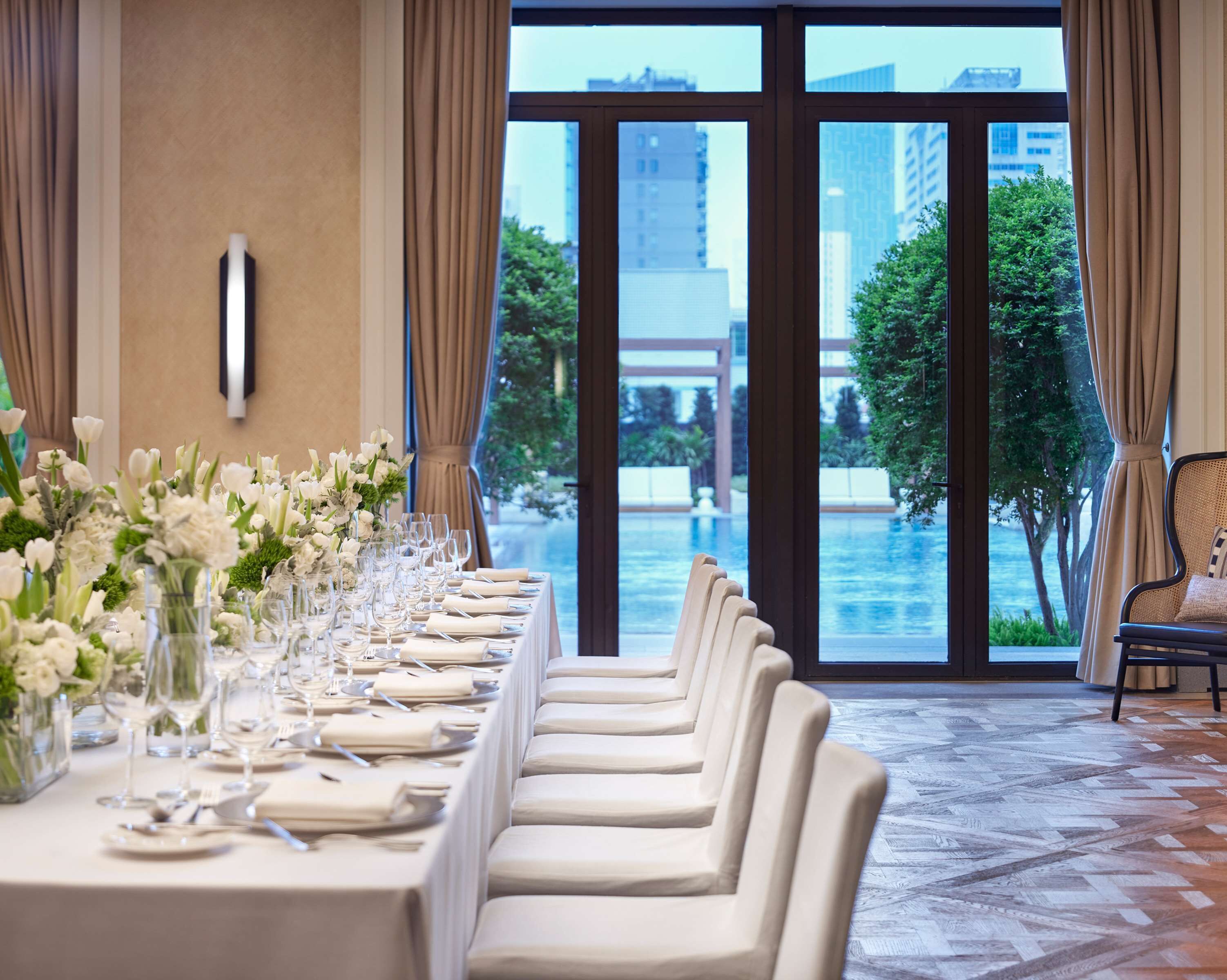 A long dining table set for a formal event with a view of a pool and buildings.