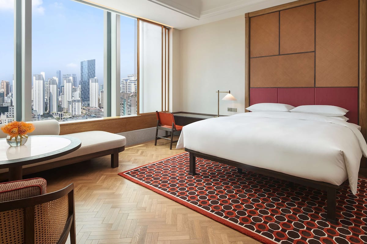 A hotel room with a large bed, a view of the cityscape, a round table with a chair, and a red patterned rug.