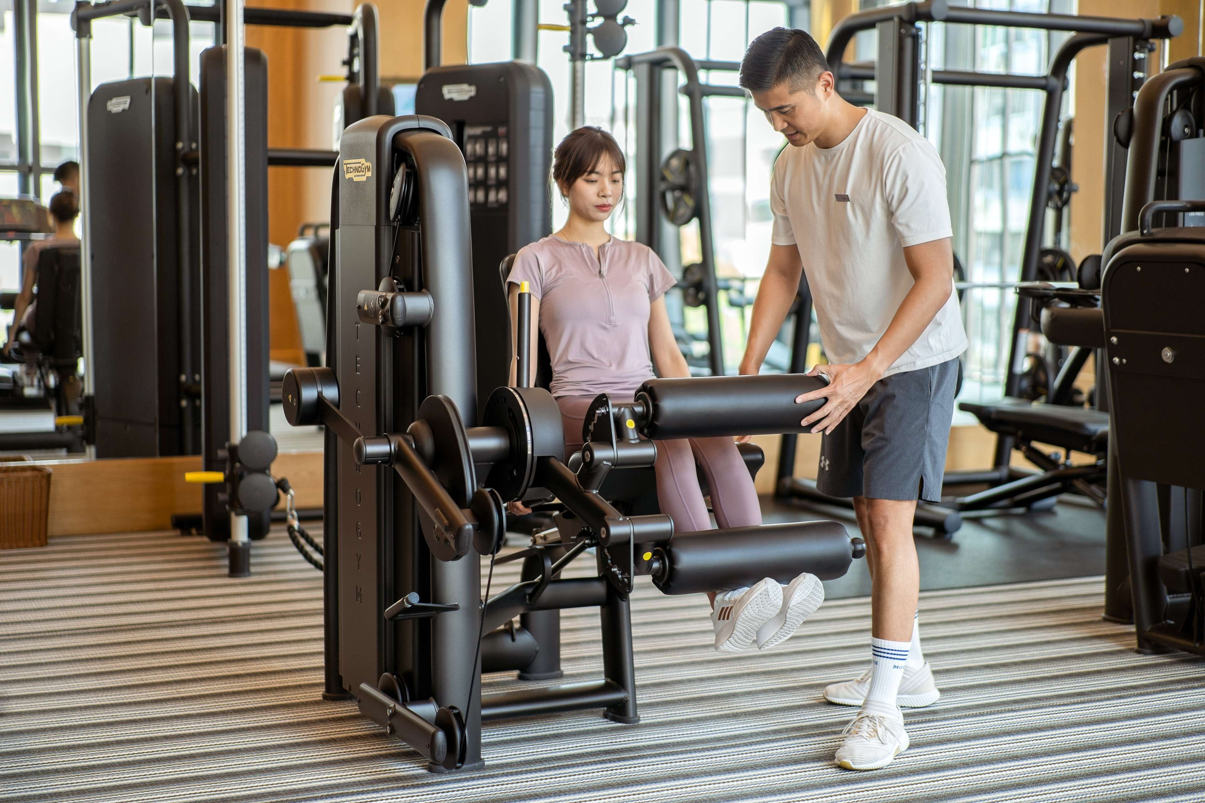 Et fitnesscenter med diverse træningsmaskiner og en person, der hjælper en anden.