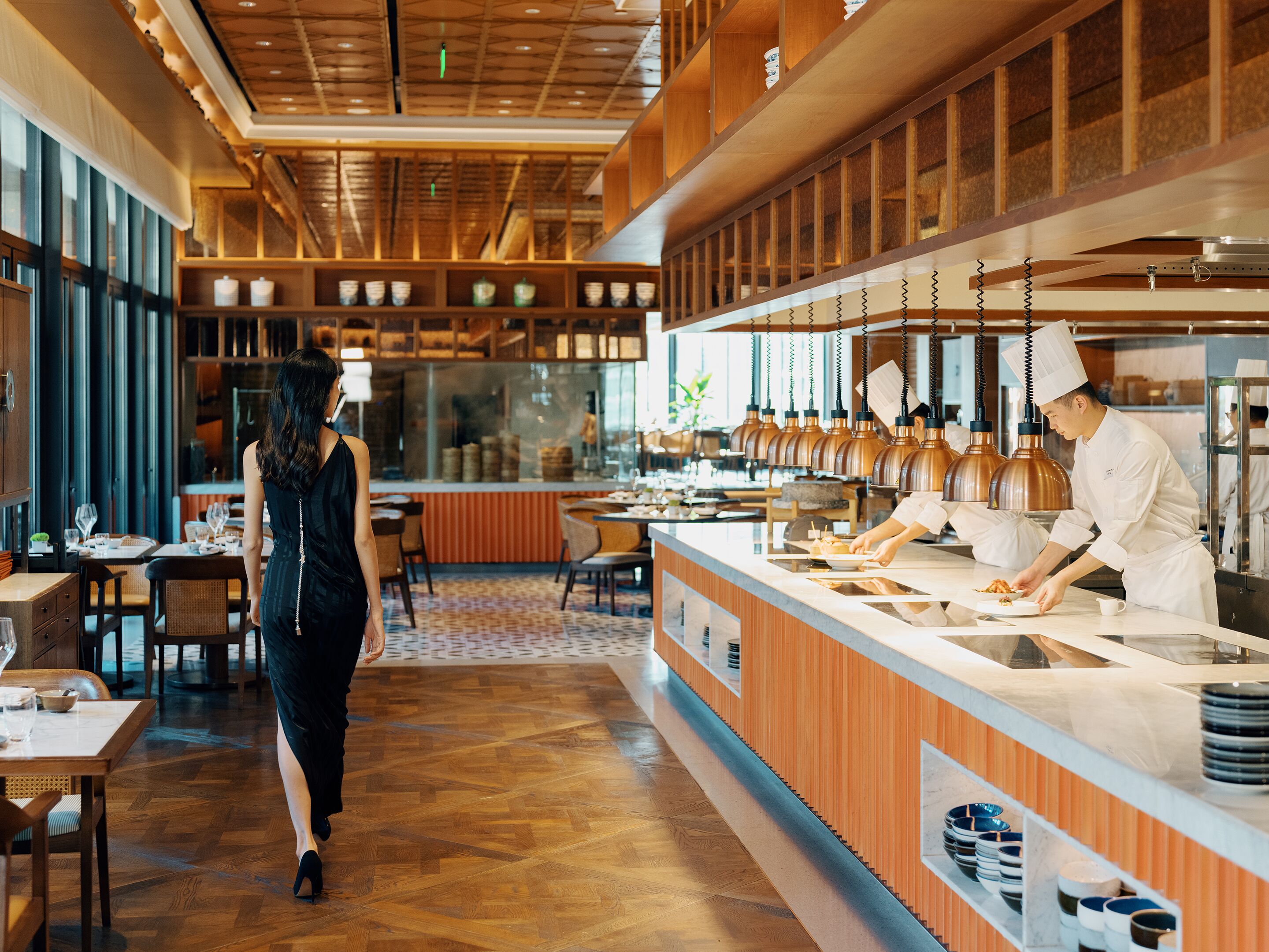 A modern restaurant interior with wooden flooring, a central serving counter, and a seating area with tables and chairs.