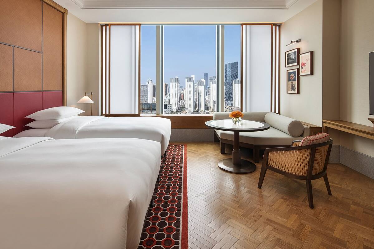 A hotel room with a large bed, a sofa, a small table with flowers, a chair, and a city view through large windows.