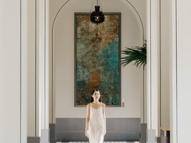 Una mujer con un vestido blanco de pie en un pasillo con un cuadro abstracto grande en la pared y una planta a la derecha.