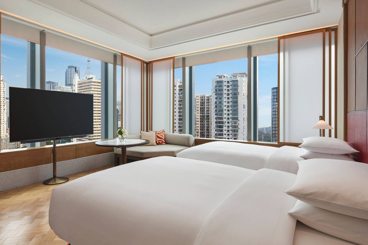 A hotel room with a large bed, a TV, a small table with a vase of flowers, and a view of tall buildings.