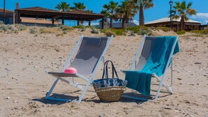 Zwei Liegestühle mit Handtüchern und einem gewebten Korb am Sandstrand, im Hintergrund Palmen und ein Gebäude.