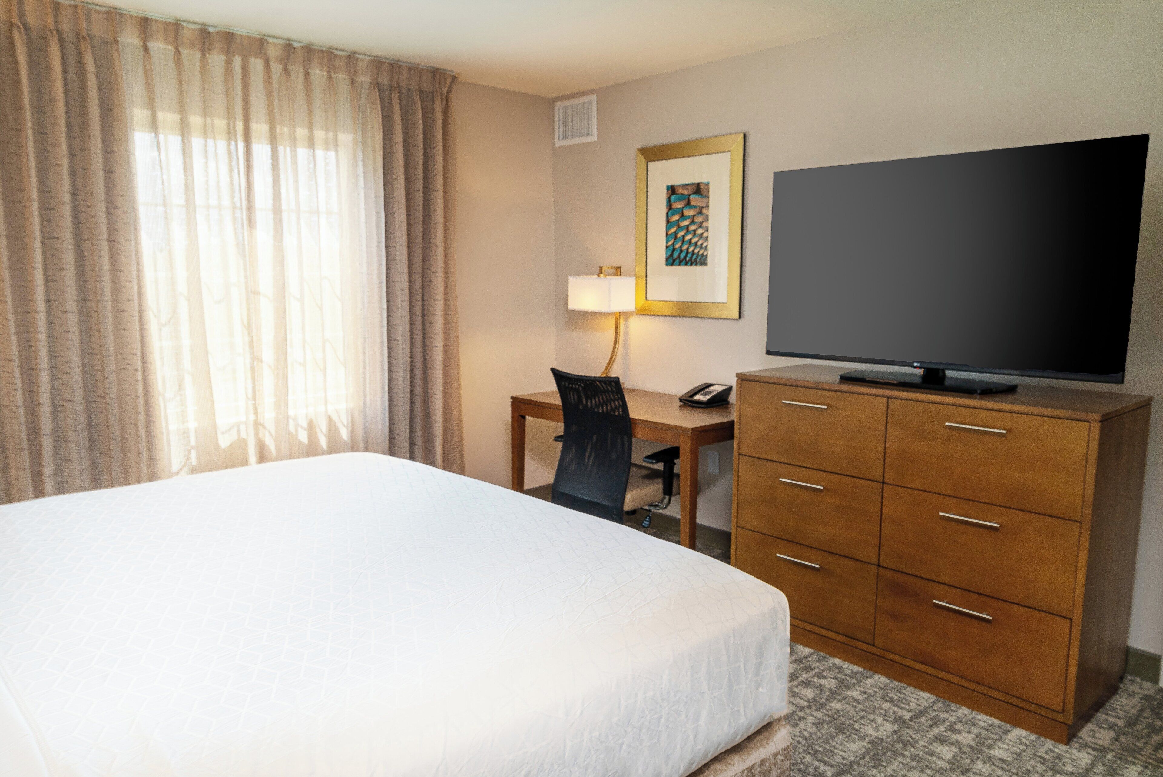 A hotel room with a bed, a desk with a chair, a television, and a wooden dresser.