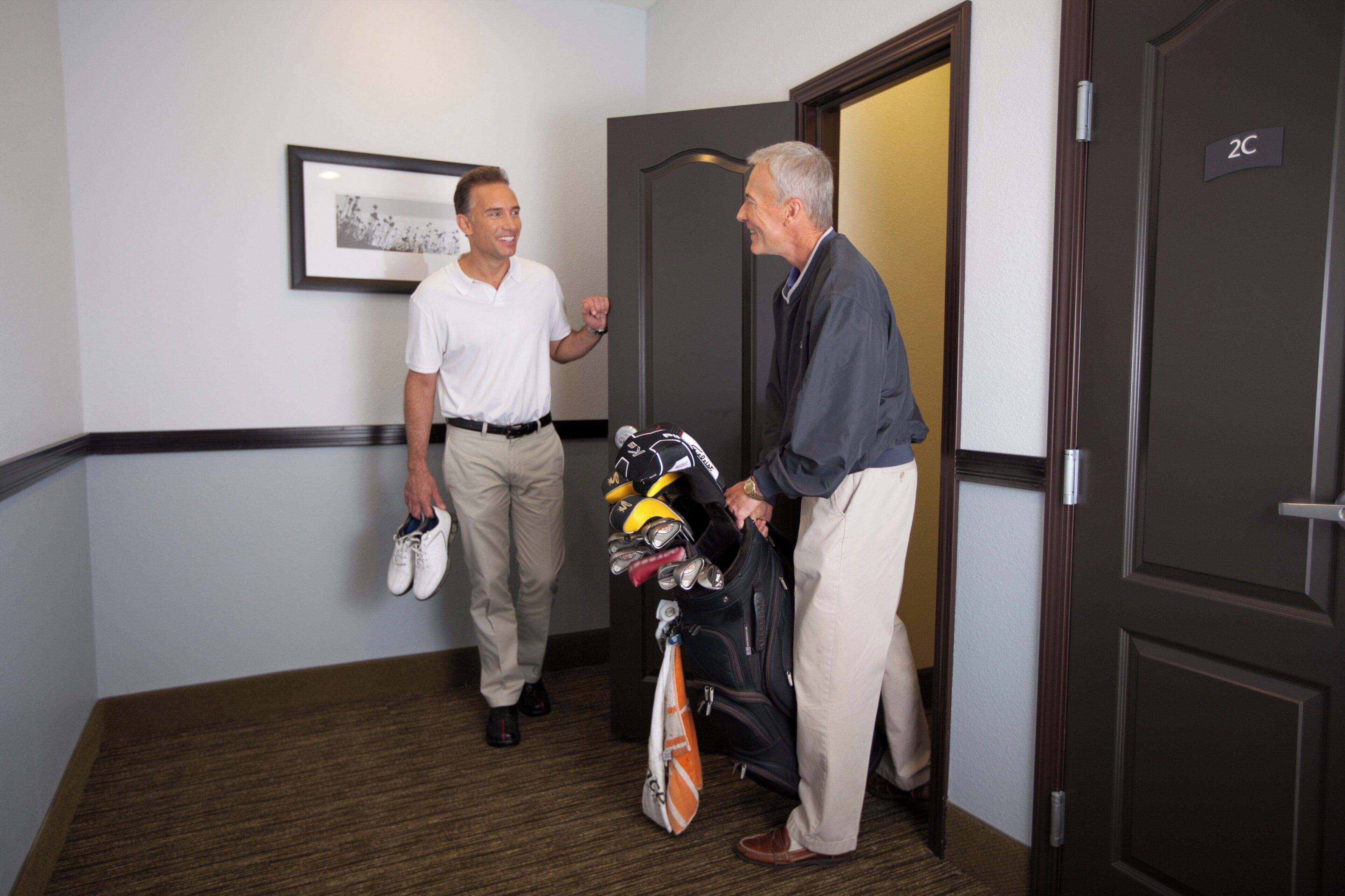 Two men in a hallway, one holding a pair of shoes and a golf bag.