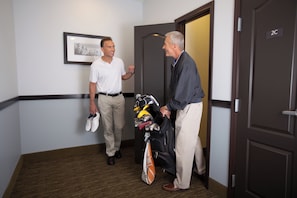 Deux hommes se trouvent dans un couloir, l’un tenant une paire de chaussures et un sac de golf.