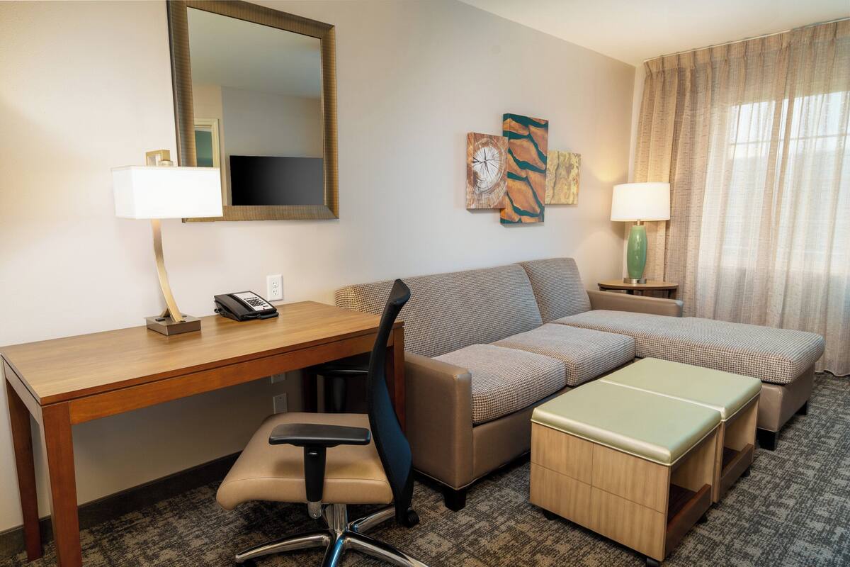 Habitación de hotel con escritorio de madera, una silla acolchada, sofá, pequeño otoman, lámpara, teléfono y cuadros en la pared.