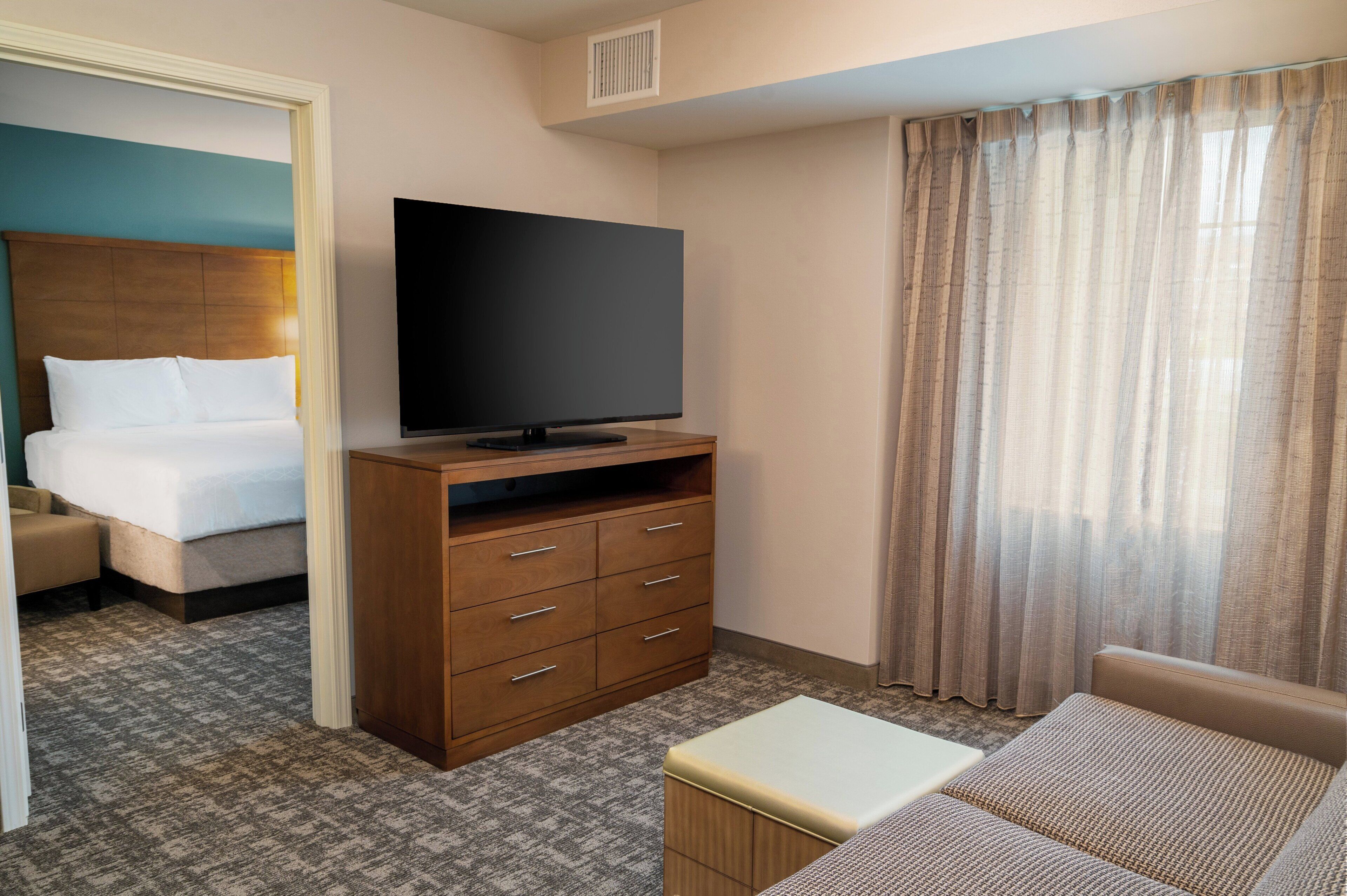A hotel room with a bed, a television on a wooden stand, a sofa, and a window with curtains.