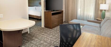 A hotel room with a bed, a TV, a desk, a chair, a sofa, and a window with curtains.