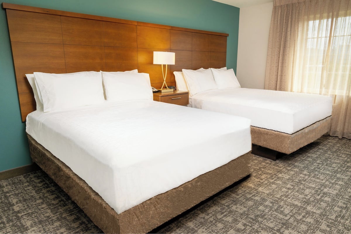 Habitación de hotel con dos camas, un cabecero de madera, una mesita de noche con lámpara y una ventana con cortinas.