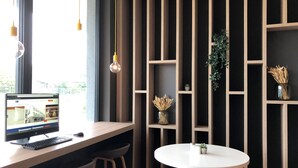 A modern workspace with a computer, two bar stools, a round table, and a vertical shelving unit with plants. - Holiday Inn Express Munich - Olympiapark by IHG (Munich)