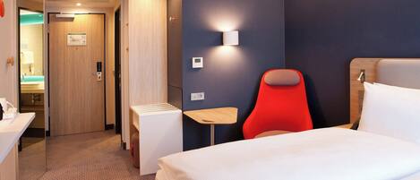 A hotel room with a bed, a red chair, a small table, a wall-mounted light, and a door leading to a bathroom.