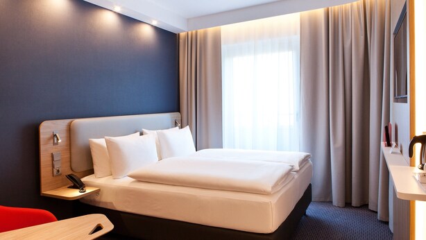 A hotel room with a bed, a desk, a chair, a nightstand, and a window with curtains. - Holiday Inn Express Munich - Olympiapark by IHG (Munich)