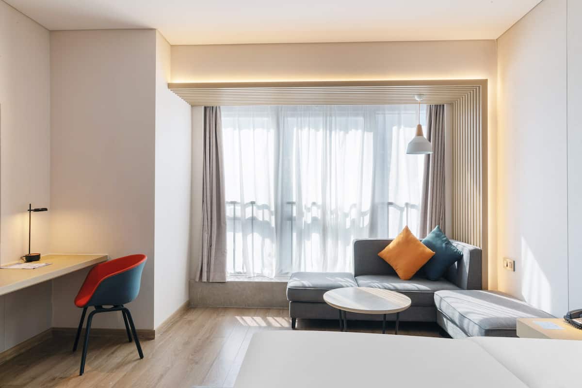 Een moderne hotelkamer met een bureau, een bank, een klein rond tafeltje en een groot raam met gordijnen.