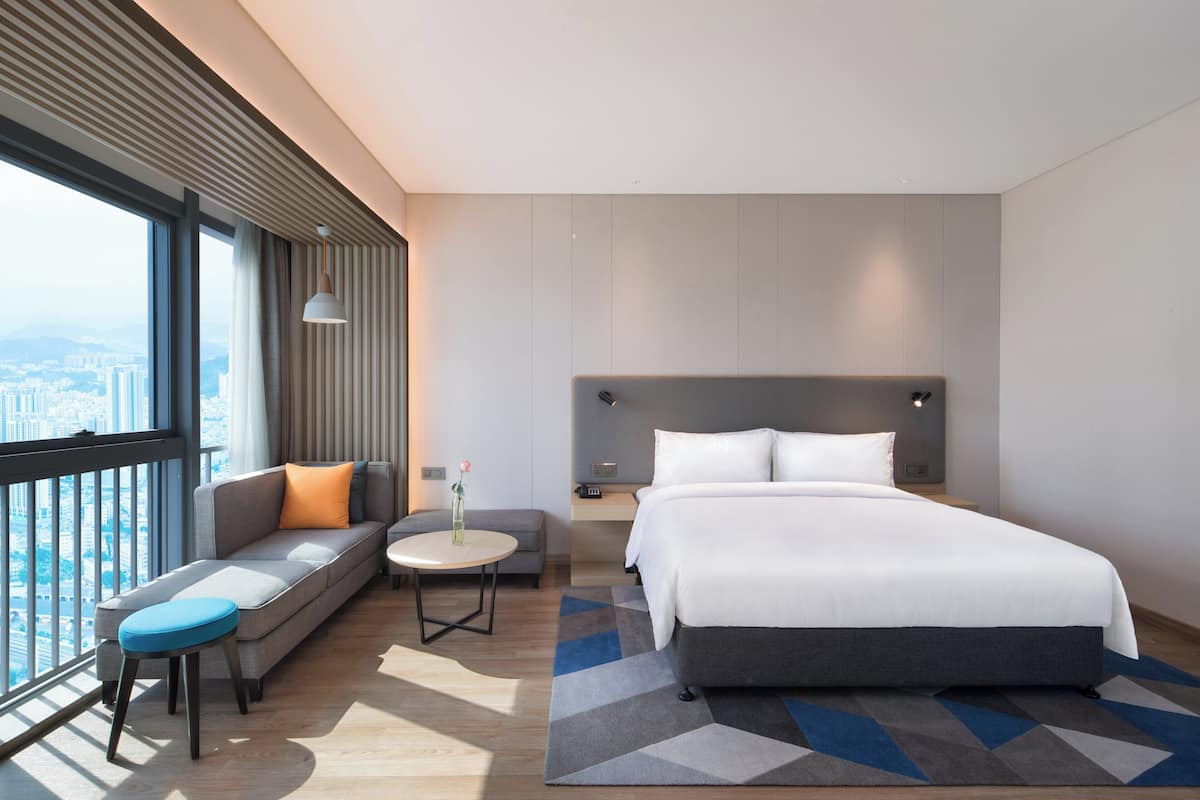 Een moderne hotelkamer met een groot bed, een bank, een klein rond tafeltje en uitzicht op de stad.