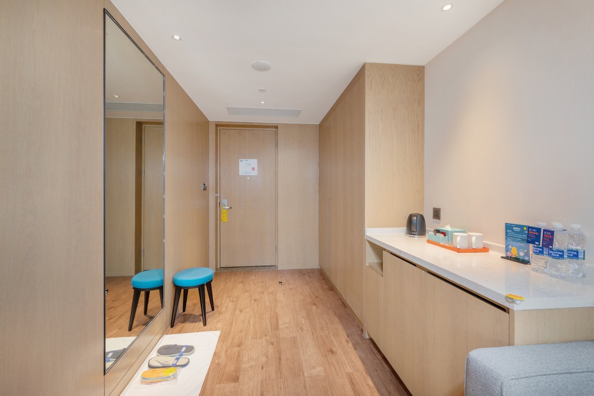 A hotel room with a wooden floor, a mirror, two blue chairs, a white counter with toiletries, and a door with a sign.