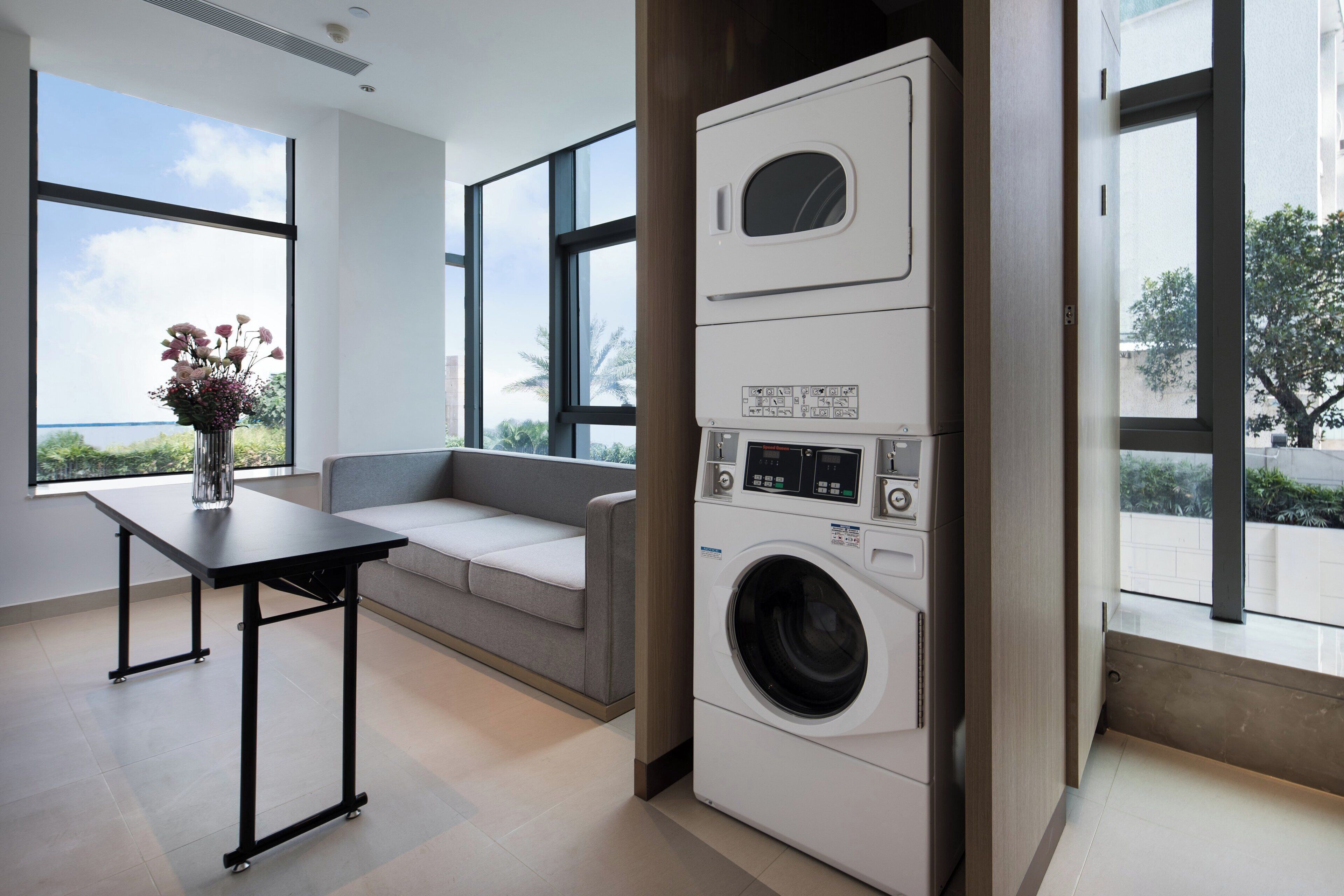 A modern laundry room with a built-in washer and dryer, a grey sofa, and large windows offering a view of palm trees.
