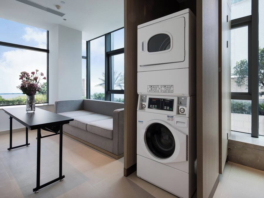 Un lavadero moderno con lavadora y secadora integradas, un sofá gris y ventanas amplias con vistas a palmeras.