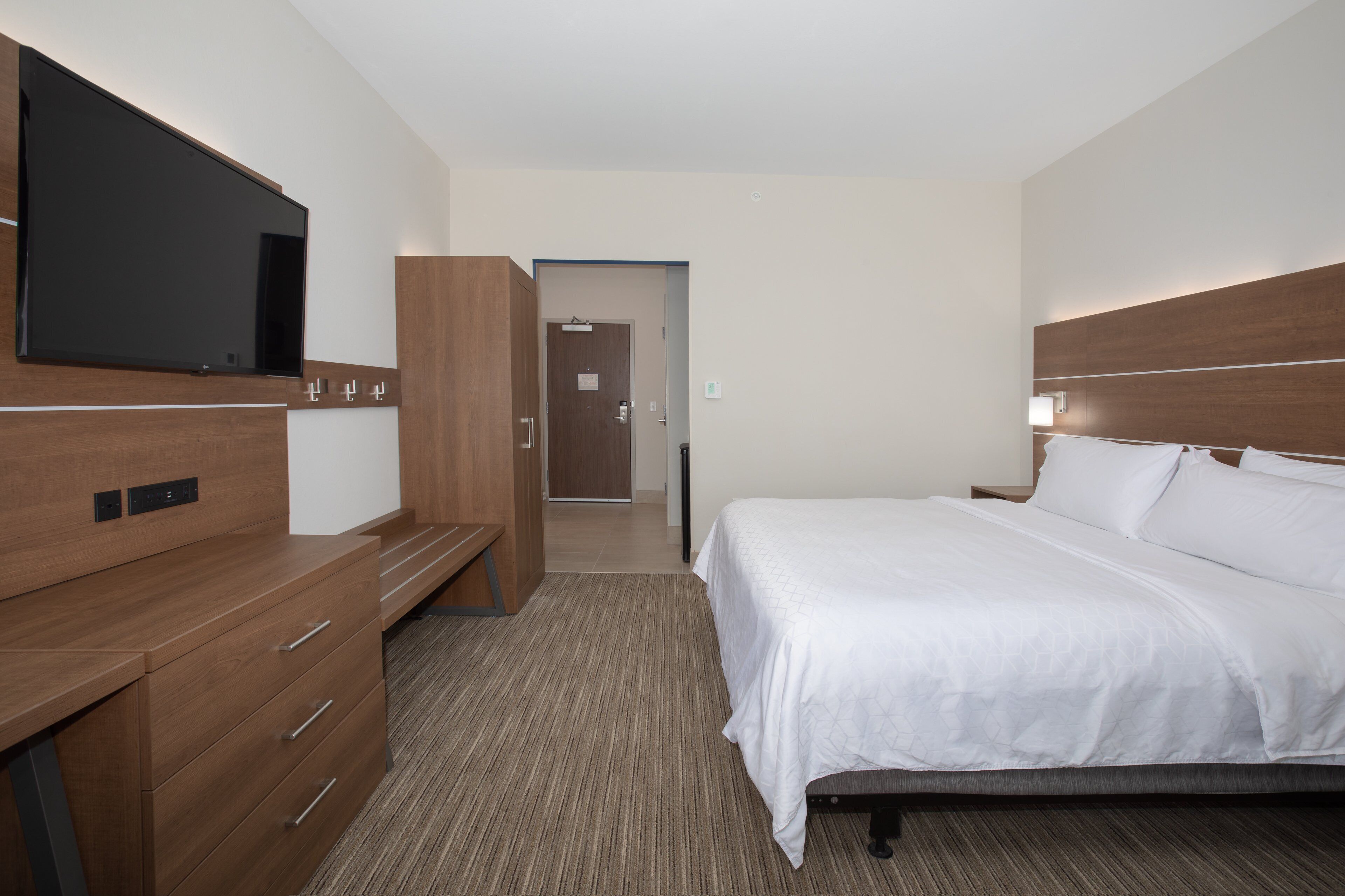 A hotel room with a bed, a TV mounted on the wall, a wooden dresser, and a door leading to another room.