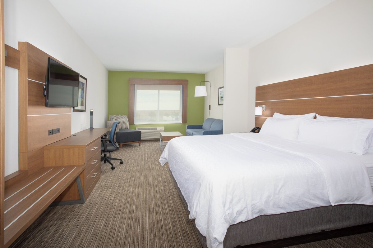 A hotel room with a large bed, a desk with a flat-screen TV, a chair, a small table, and a window with a view.