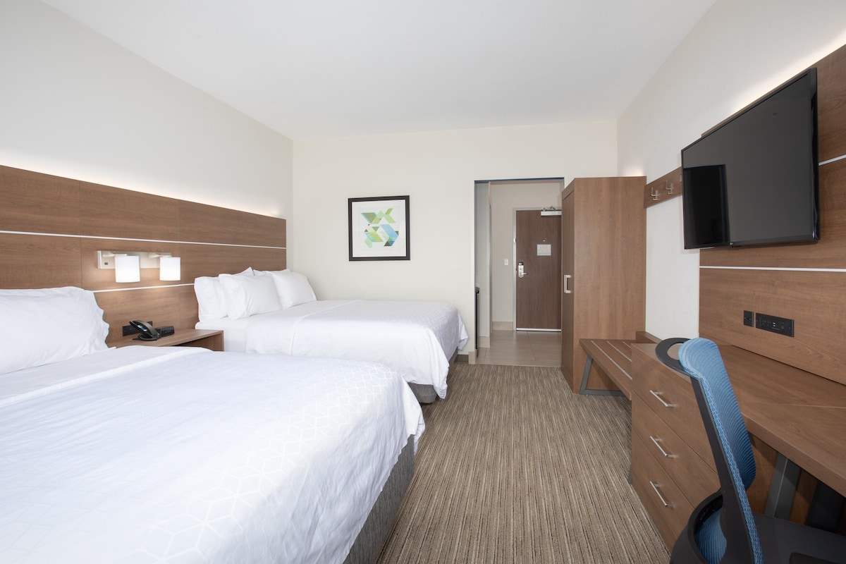 Quarto de hotel com duas camas, uma secretária com cadeira, televisão de tela plana, guarda-roupa de madeira e um quadro pendurado na parede.