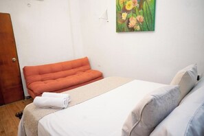 Une chambre à coucher avec un lit, un coussin orange, du linge de lit blanc et un tableau représentant des fleurs accroché au mur.