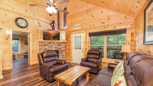 A log cabin interior with leather recliners, a wooden coffee table, a stone fireplace, and a large window offering a view of the outdoors. - Free Tickets | Hot Tub, Pool Table, Media Room, Easy Access Crazy Like a Fox! (Pigeon Forge)