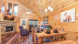 A rustic cabin interior with a stone fireplace, wooden dining table, leather sofa, and a ceiling fan. - Free Tickets | Hot Tub, Pool Table, Media Room, Easy Access Crazy Like a Fox! (Pigeon Forge)